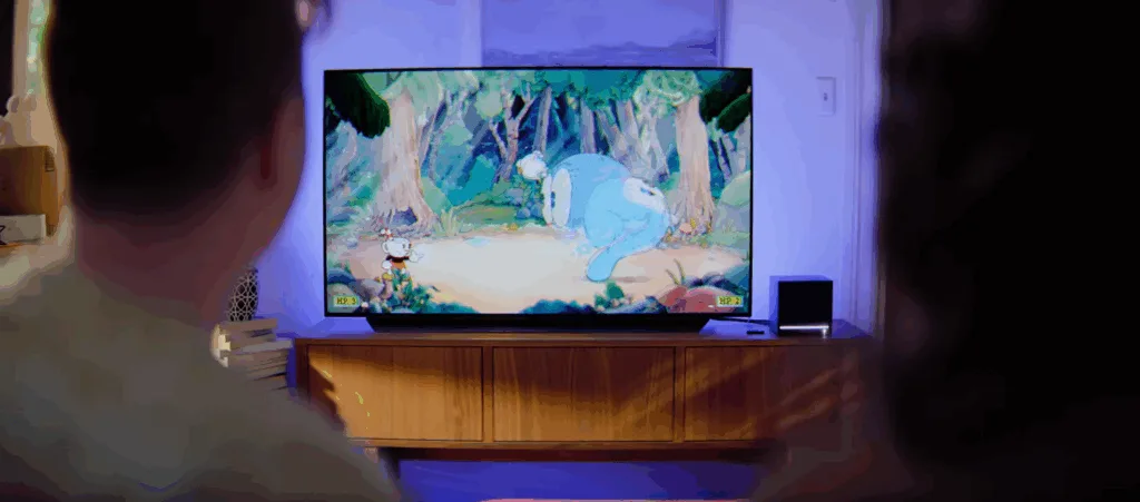 A picture of two people sitting on a couch in the living room, using a Steam Machine to play Cuphead on a big screen TV.