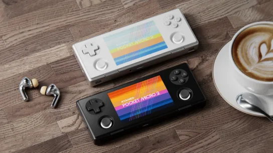 A promotional picture of the Ayaneo Pocket Micro 2, showing both the White and Black colors sitting on a desk with wireless earbuds on the left, and a cup of coffee on the right.